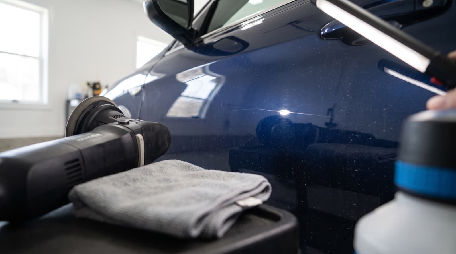 Close-up of a light scratch on a dark car door, with a machine polisher and a folded microfiber towel resting beside it