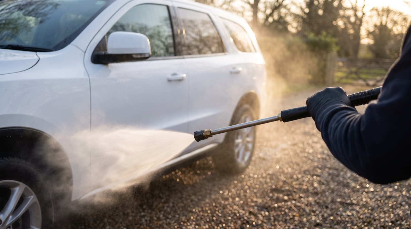 A person rinses a silver sedan in a driveway with a pressure washer wand, standing well back and spraying a wide, flat fan of water from the roof down