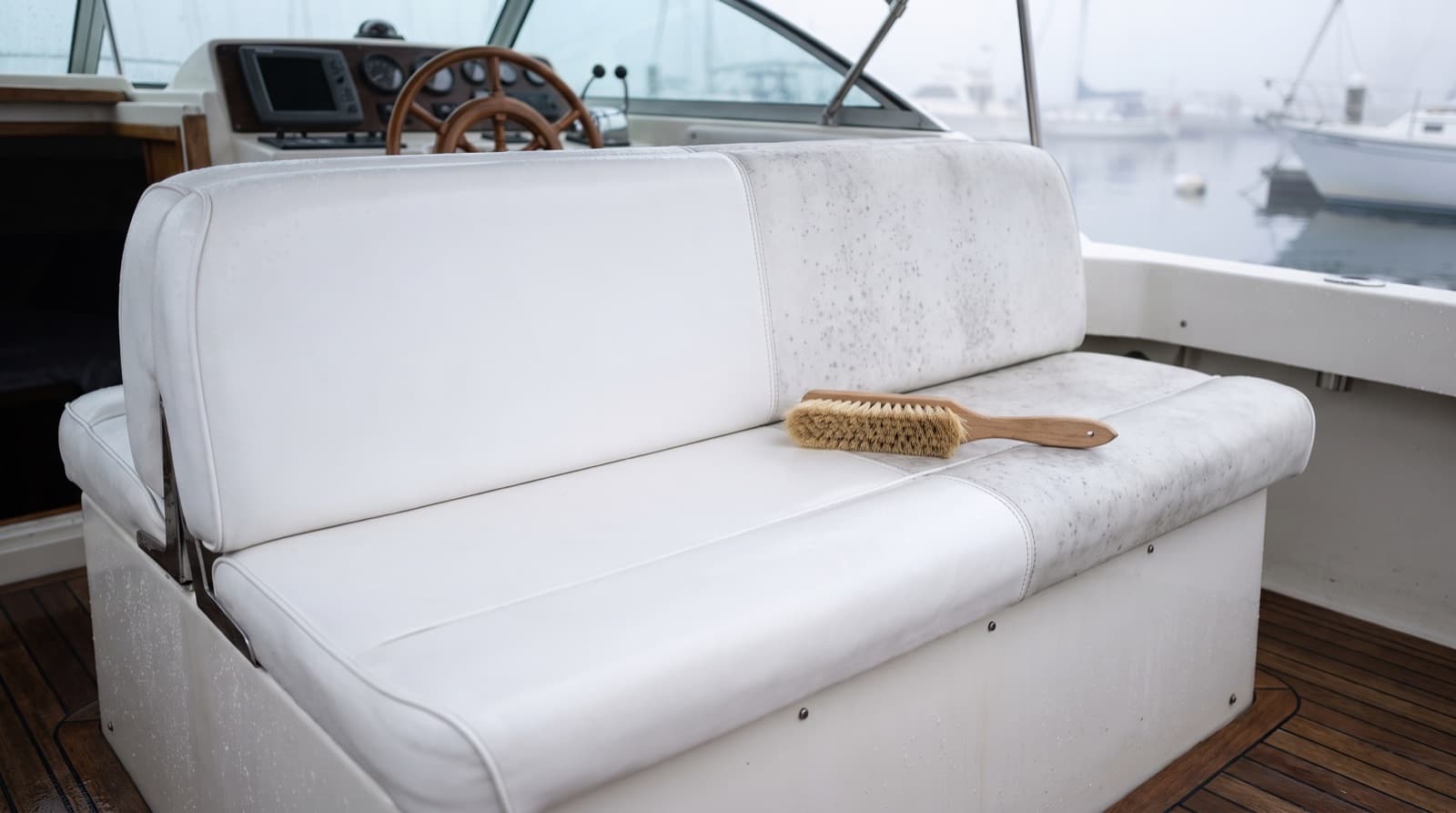 Gloved hands scrubbing a white marine vinyl boat seat with a soft brush under a shaded boat canopy, with a spray bottle and folded microfiber towels beside it