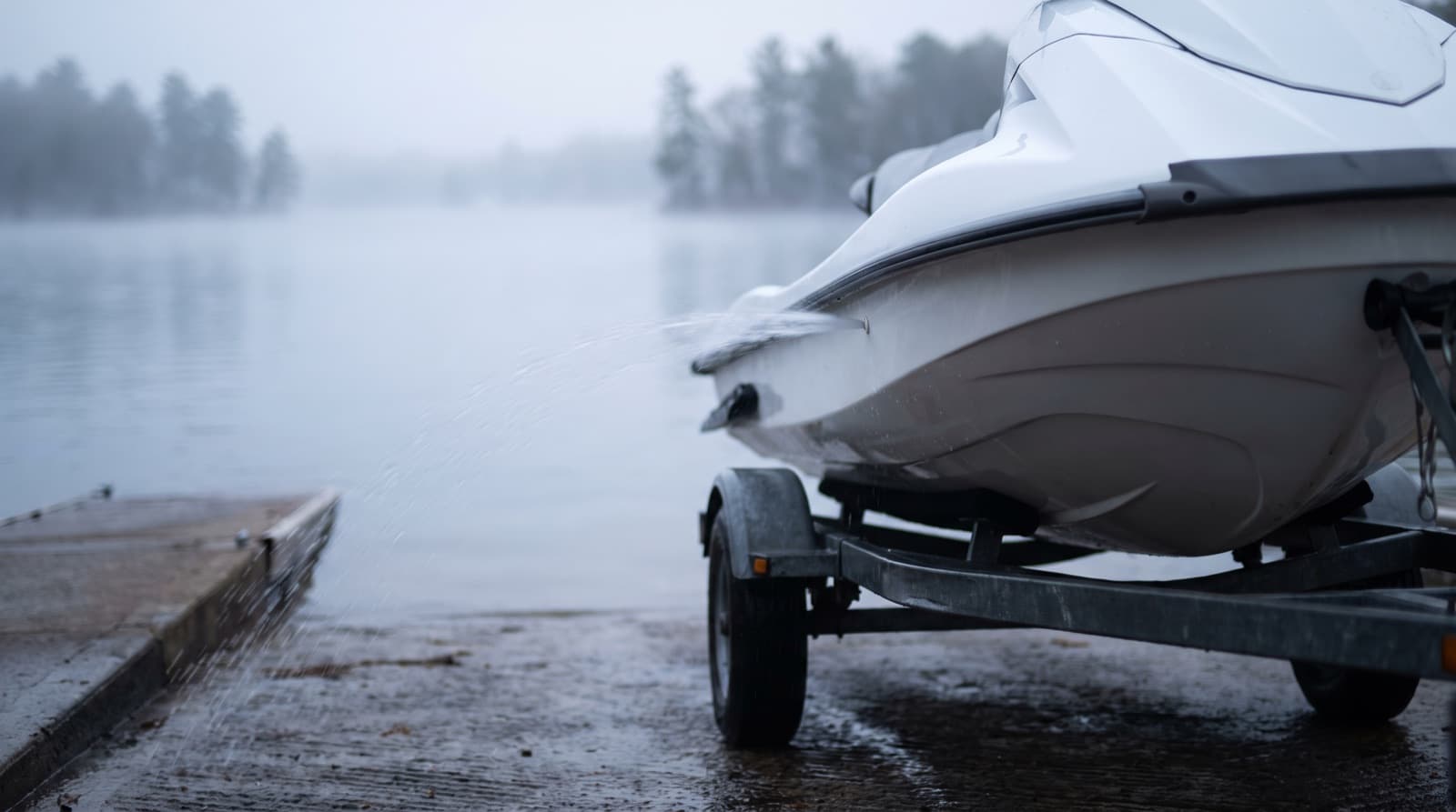 A white jet ski on its trailer in a driveway being rinsed with a garden hose, with dried salt spots on the hull and a gray vinyl seat