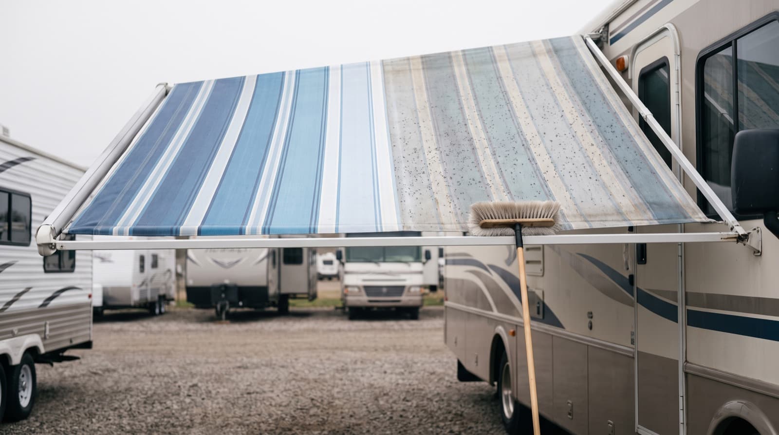 A striped RV patio awning rolled all the way out beside a white travel trailer in a driveway, with a soft brush on a long pole and a garden hose on the ground below it
