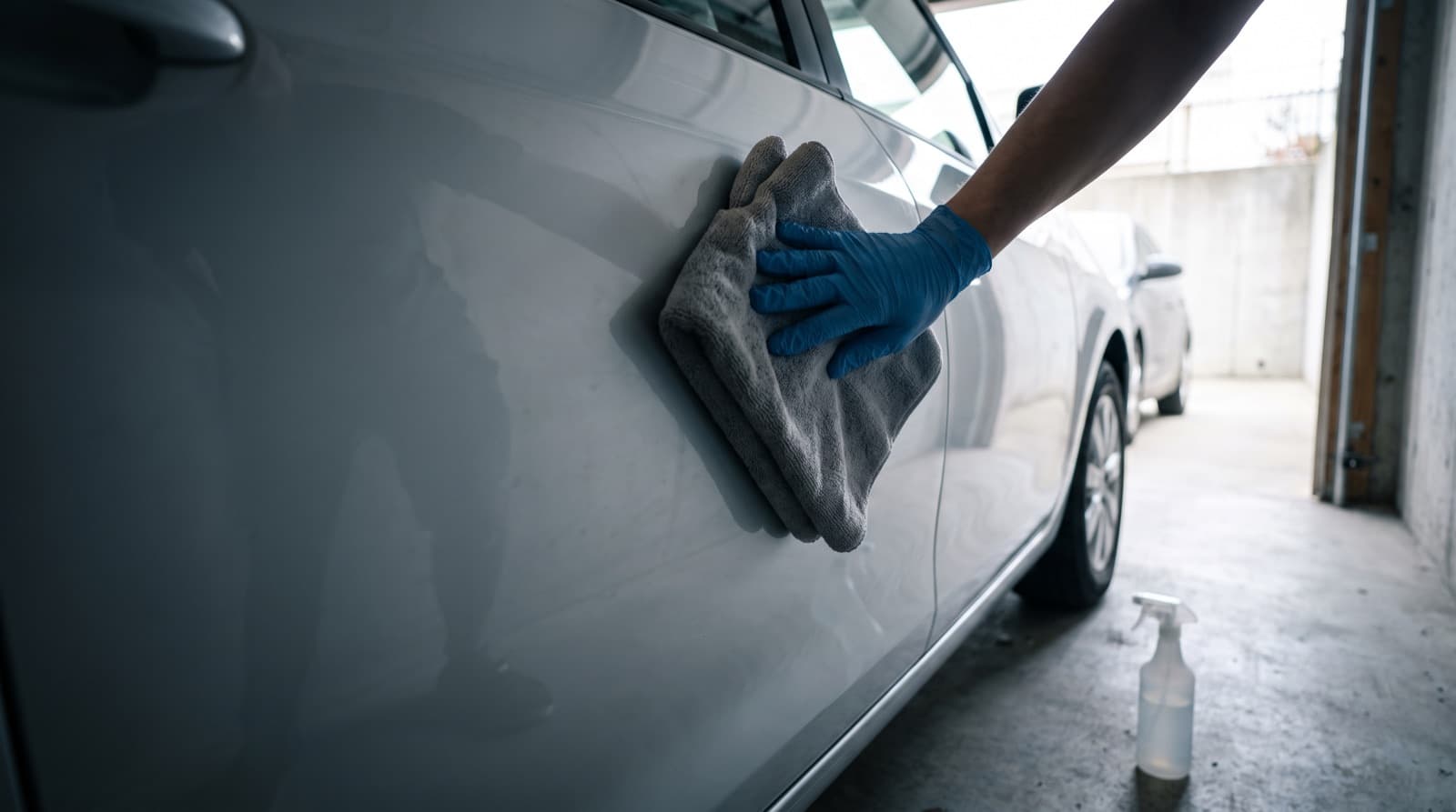 A gloved hand wiping light dust off a dark gray car hood with a folded blue microfiber towel, a spray bottle and a stack of clean towels nearby in an apartment garage