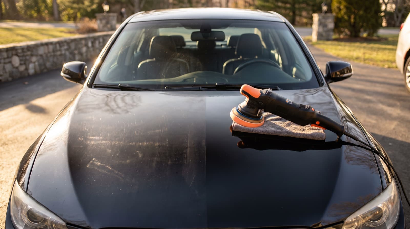 A detailer holds a dual action polisher flat on the hood of a dark blue car, with blue painter's tape on the trim and a bright work light beside it