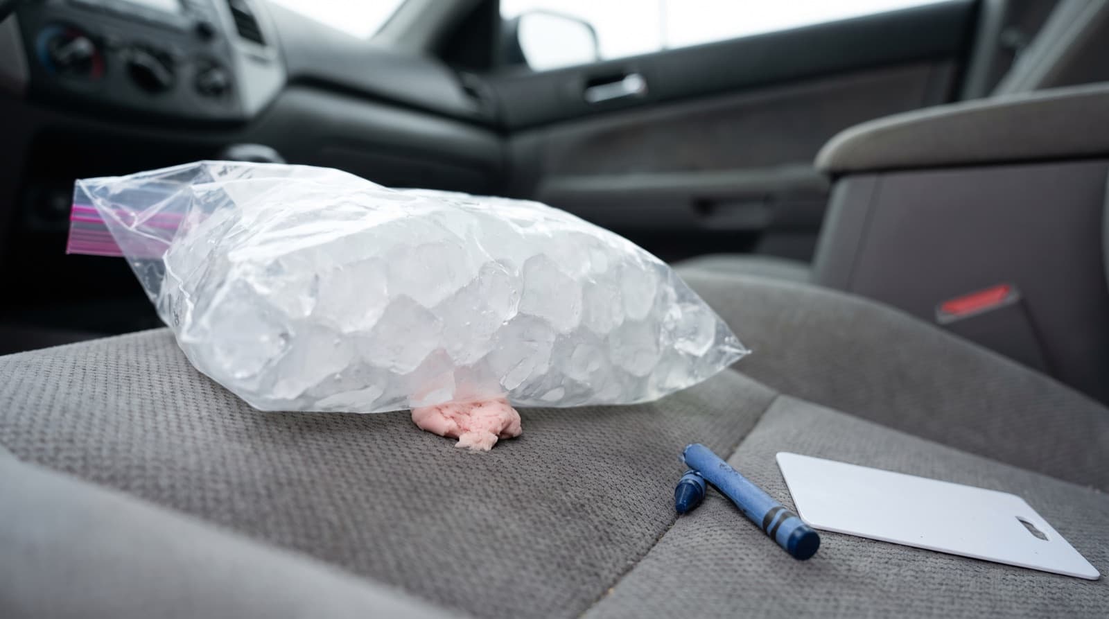 A bag of ice resting on a piece of chewing gum stuck to a gray cloth car seat, with an old plastic gift card ready to lift it off