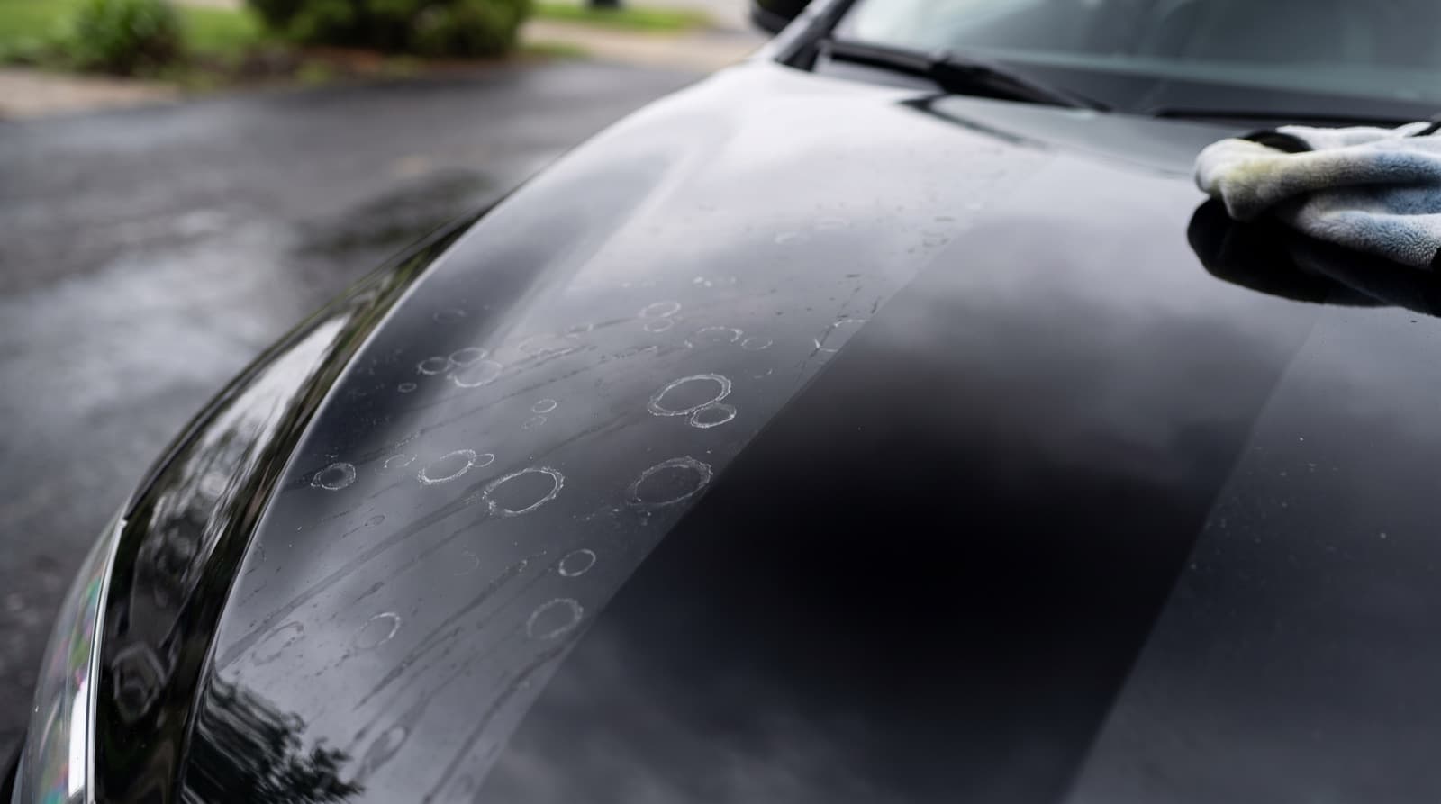 Close-up of round water spots dried on the hood of a dark ceramic coated car, with a microfiber towel and a spray bottle resting nearby
